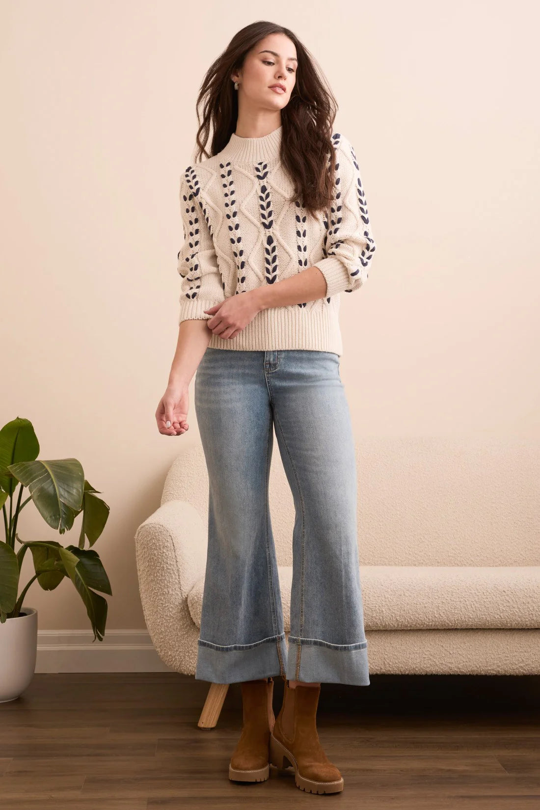 Woman wearing a patterned sweater, jeans, and boots in a room with a plant and sofa.