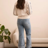 Woman wearing a white cable knit sweater and blue jeans standing in a room with a plant and sofa.