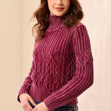 A woman wearing a textured, cable-knit mock neck sweater in a deep berry purple, paired with blue jeans, standing against a neutral beige wall.