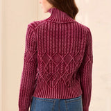 A woman shown from the back wearing a burgundy cable-knit mock neck sweater and blue jeans, set against a neutral beige background.
