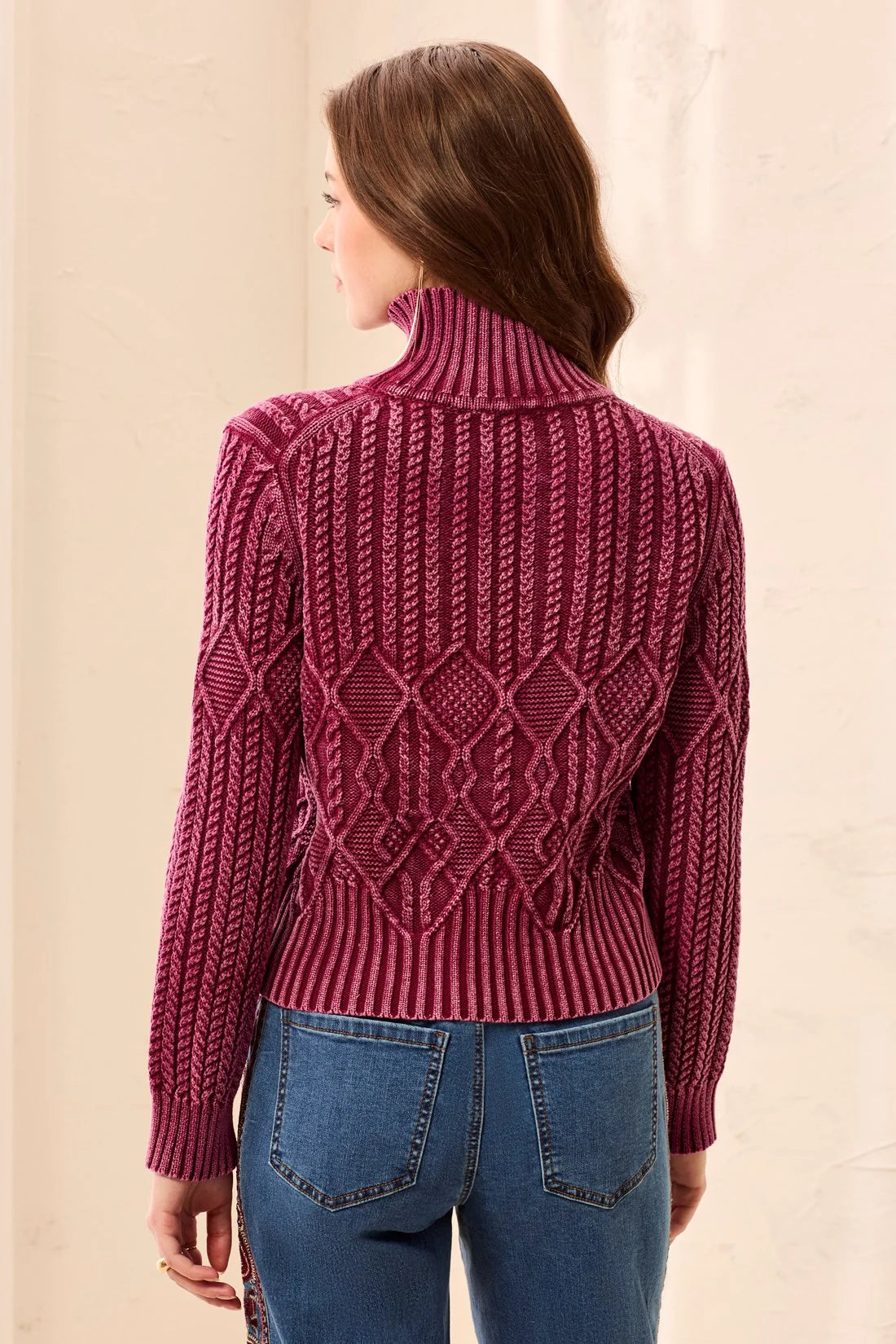 A woman shown from the back wearing a burgundy cable-knit mock neck sweater and blue jeans, set against a neutral beige background.