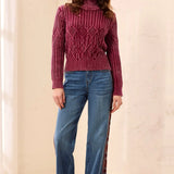 A woman modeling a burgundy cable-knit mock neck sweater paired with wide-leg blue jeans featuring a burgundy side stripe and burgundy shoes, set against a warm beige textured wall.