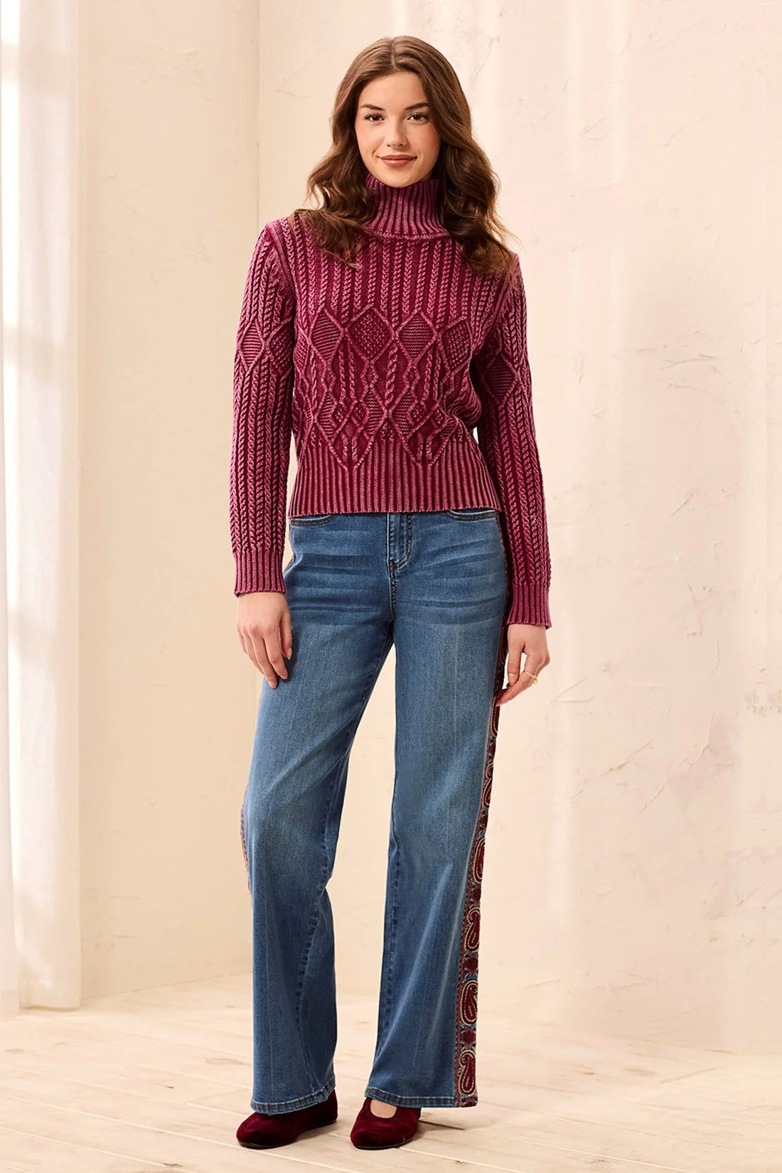 A woman modeling a burgundy cable-knit mock neck sweater paired with wide-leg blue jeans featuring a burgundy side stripe and burgundy shoes, set against a warm beige textured wall.