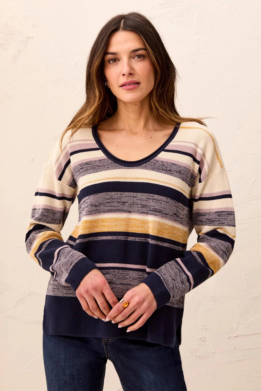 A woman wearing a long-sleeve knit sweater with horizontal stripes in cream, navy, gray, and mustard yellow, paired with dark blue jeans, standing against a textured off-white wall.