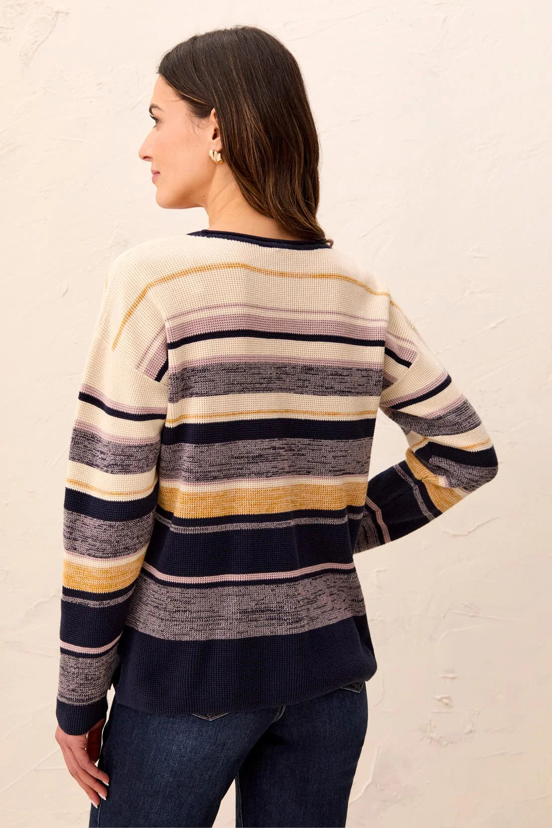 A woman shown from the back wearing a long-sleeve knit sweater with horizontal stripes in cream, navy, gray, and mustard yellow, paired with dark blue jeans against a textured off-white wall.