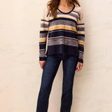 A woman modeling a long-sleeve knit sweater with horizontal stripes in navy, gray, beige, and mustard yellow, paired with dark blue jeans and navy sneakers, standing against a textured off-white wall.