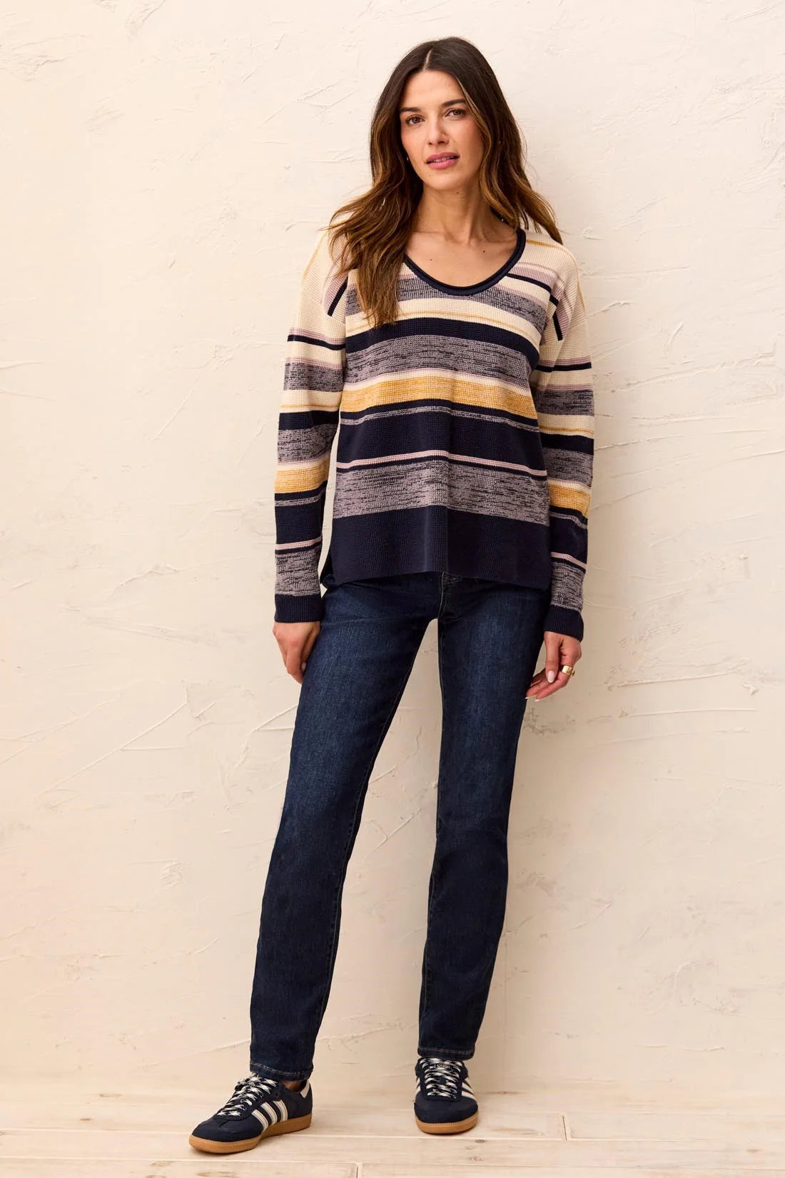 A woman modeling a long-sleeve knit sweater with horizontal stripes in navy, gray, beige, and mustard yellow, paired with dark blue jeans and navy sneakers, standing against a textured off-white wall.