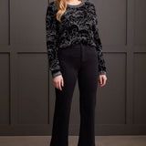 Woman wearing a black patterned sweater and black pants against a gray wall.
