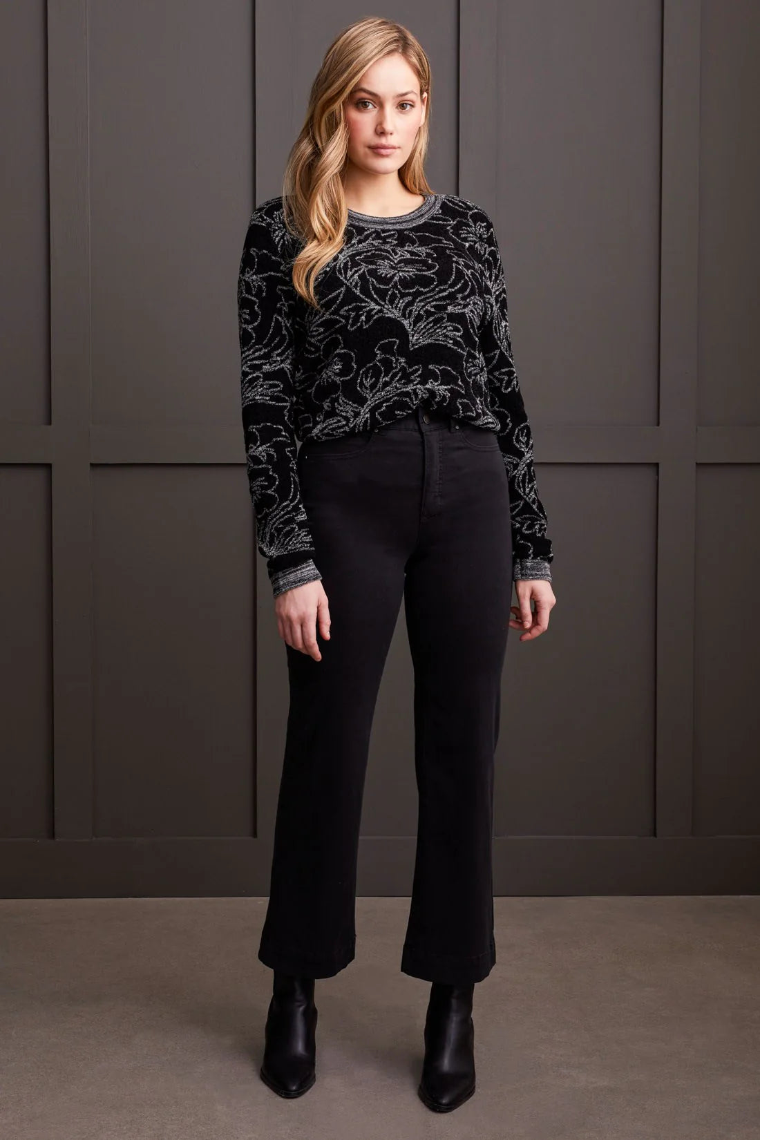 Woman wearing a black patterned sweater and black pants against a gray wall.
