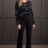 Woman wearing a black patterned sweater and black pants against a gray wall.