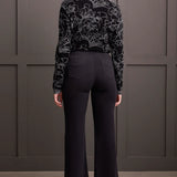Woman wearing a black patterned top and black pants against a gray wall.