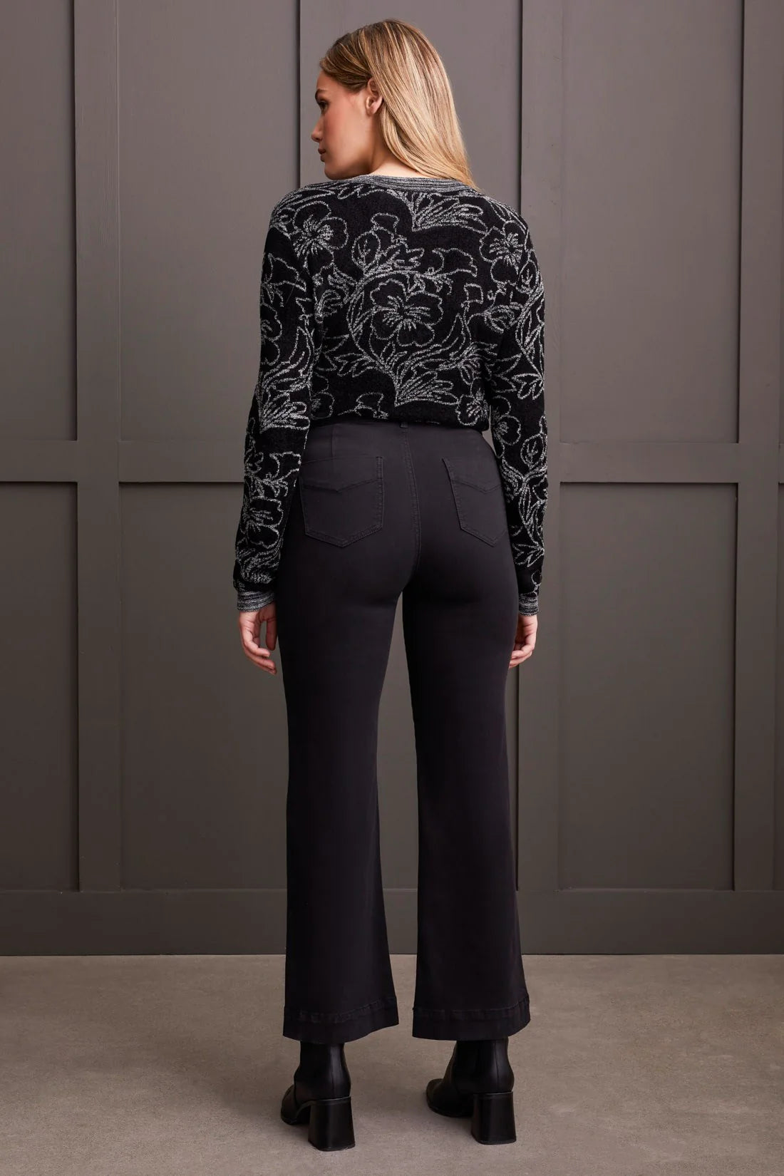 Woman wearing a black patterned top and black pants against a gray wall.