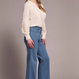 Woman wearing a cream blouse, blue jeans, and brown boots against a plain background