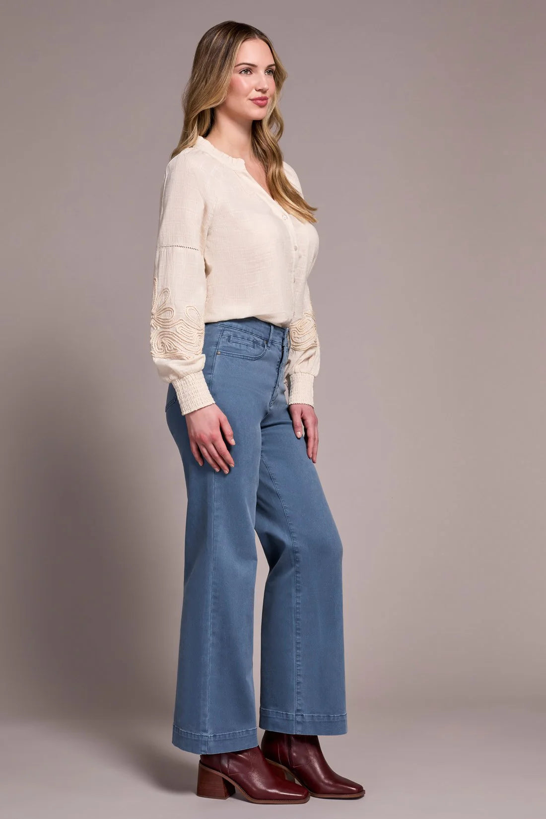 Woman wearing a cream blouse, blue jeans, and brown boots against a plain background
