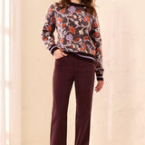 A woman modeling a long-sleeve crewneck sweater with a multicolor floral pattern in purple, orange, and green on a gray base, paired with dark burgundy pants and beige sneakers, set against a warm cream-colored indoor background.