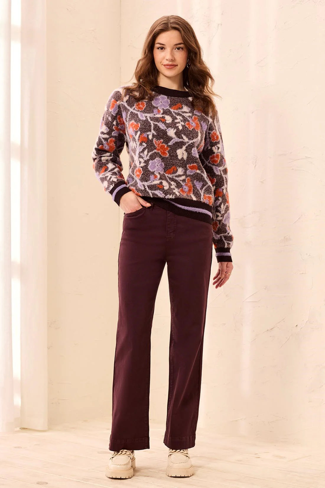 A woman modeling a long-sleeve crewneck sweater with a multicolor floral pattern in purple, orange, and green on a gray base, paired with dark burgundy pants and beige sneakers, set against a warm cream-colored indoor background.