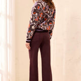 A woman stands in a bright indoor setting wearing a long-sleeve sweater with an abstract floral pattern in orange, purple, and black, paired with dark plum wide-leg pants and cream-colored shoes.