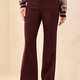 A person wearing dark burgundy flare jeans, a patterned knit sweater, and cream lace-up shoes, standing against a light beige textured wall.