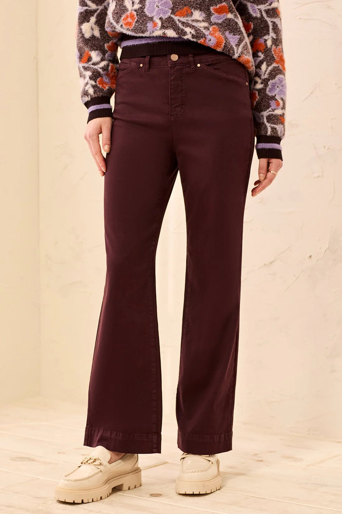 A person wearing dark burgundy flare jeans, a patterned knit sweater, and cream lace-up shoes, standing against a light beige textured wall.