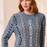 A woman with red hair wearing a dusty blue cable-knit sweater with vertical embroidered leaf and wheat-like patterns in navy, cream, and mustard yellow, set against a soft beige background.