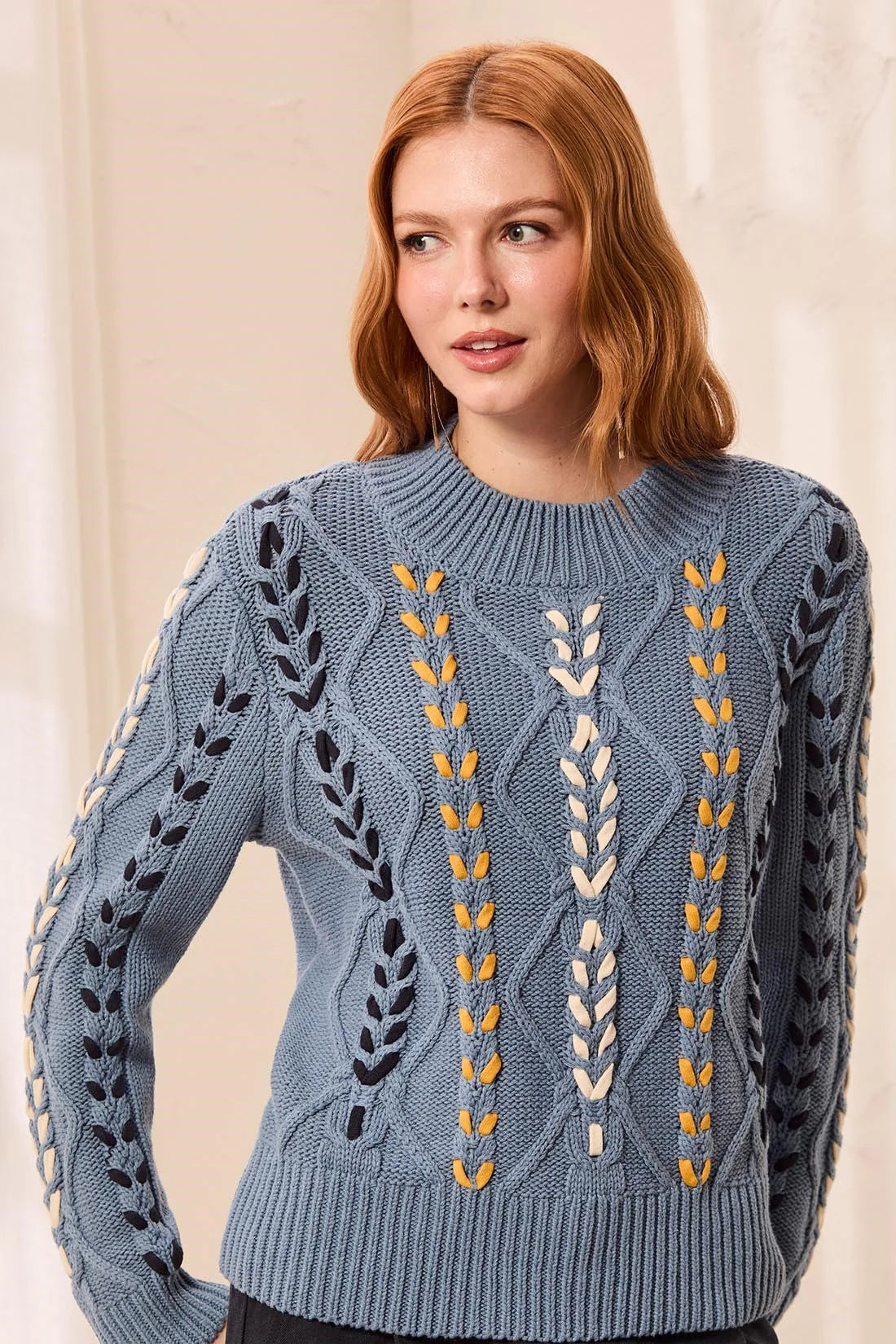 A woman with red hair wearing a dusty blue cable-knit sweater with vertical embroidered leaf and wheat-like patterns in navy, cream, and mustard yellow, set against a soft beige background.