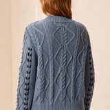 A person shown from the back wearing a textured cable-knit sweater in a dusty slate blue color, paired with dark pants, against a neutral beige wall.