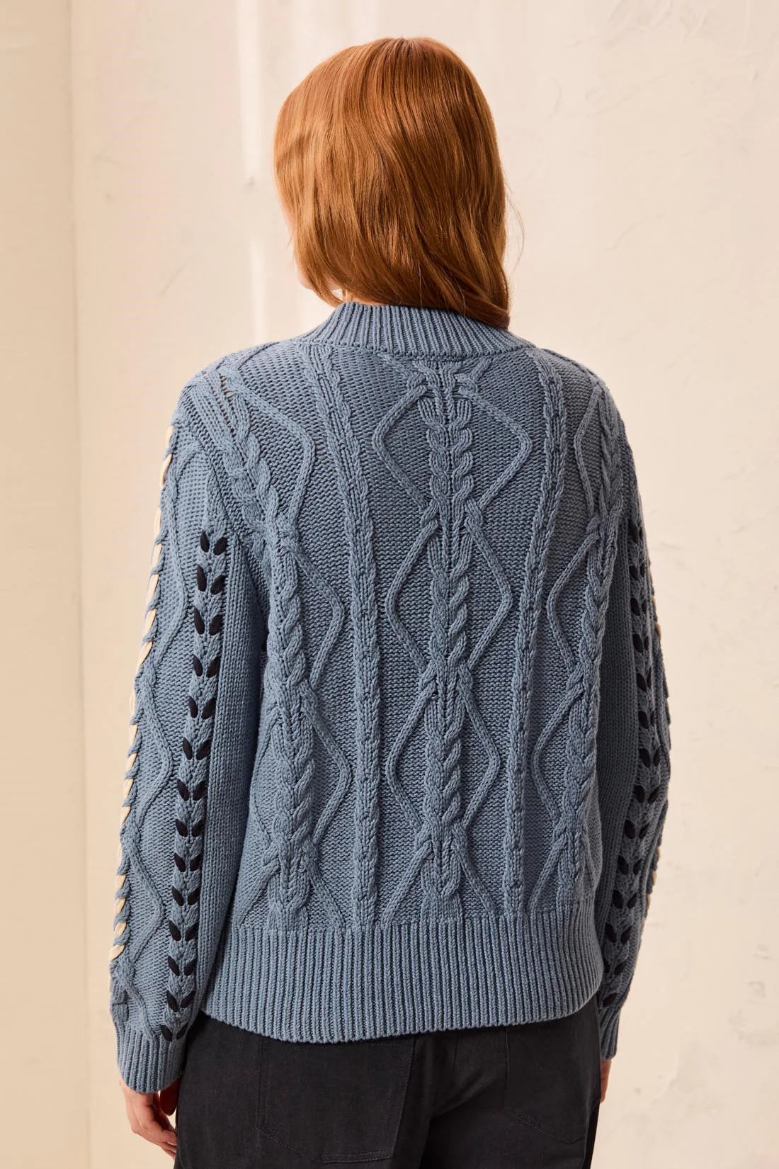 A person shown from the back wearing a textured cable-knit sweater in a dusty slate blue color, paired with dark pants, against a neutral beige wall.