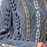 A close-up of a textured blue cable-knit sweater with vertical embroidered leaf and wheat-like patterns in black, mustard yellow, and cream, worn by a model.