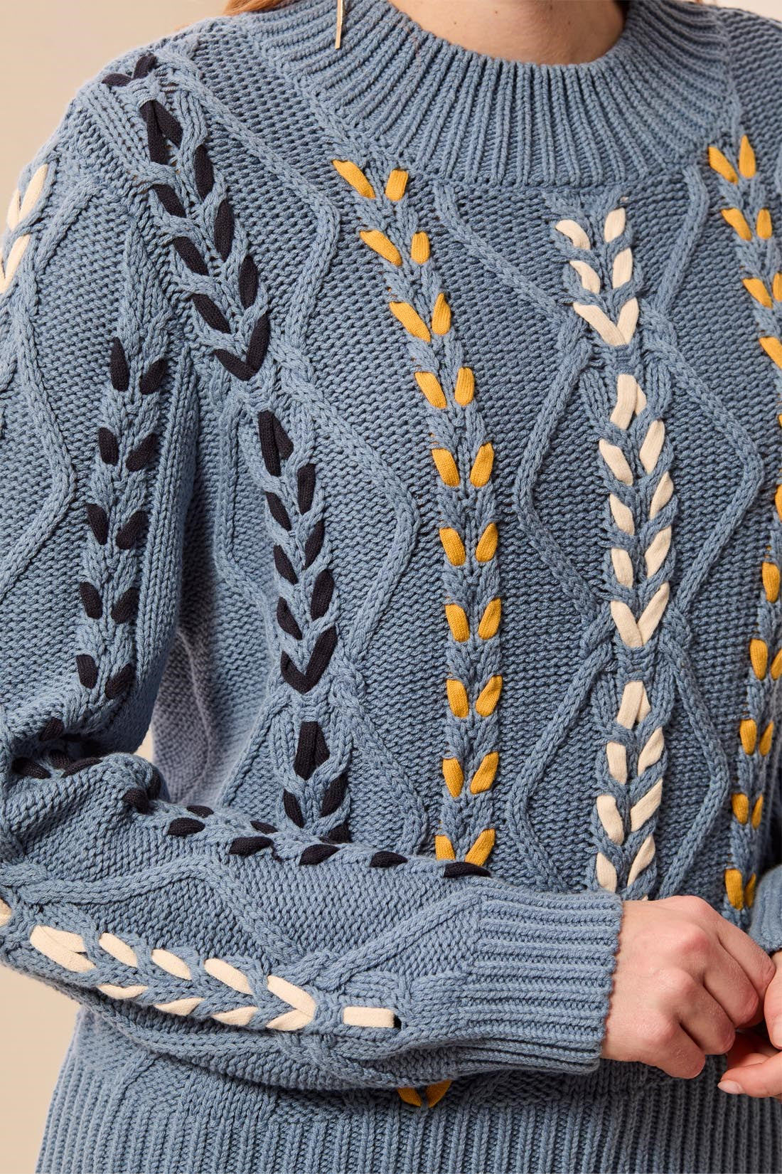 A close-up of a textured blue cable-knit sweater with vertical embroidered leaf and wheat-like patterns in black, mustard yellow, and cream, worn by a model.