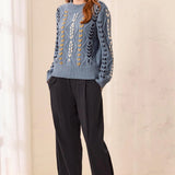 A woman modeling a light blue cable-knit sweater with white and yellow geometric patterns, paired with dark charcoal wide-leg trousers and brown suede loafers, standing in a brightly lit room with a cream wall and window curtains.