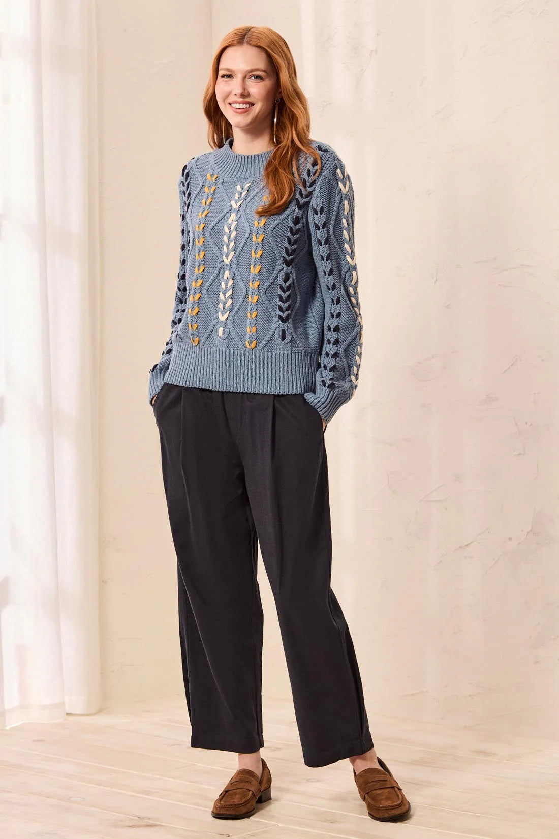 A woman modeling a light blue cable-knit sweater with white and yellow geometric patterns, paired with dark charcoal wide-leg trousers and brown suede loafers, standing in a brightly lit room with a cream wall and window curtains.