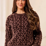 A woman wearing a brown and black cable-knit crewneck sweater, standing against a light beige wall.