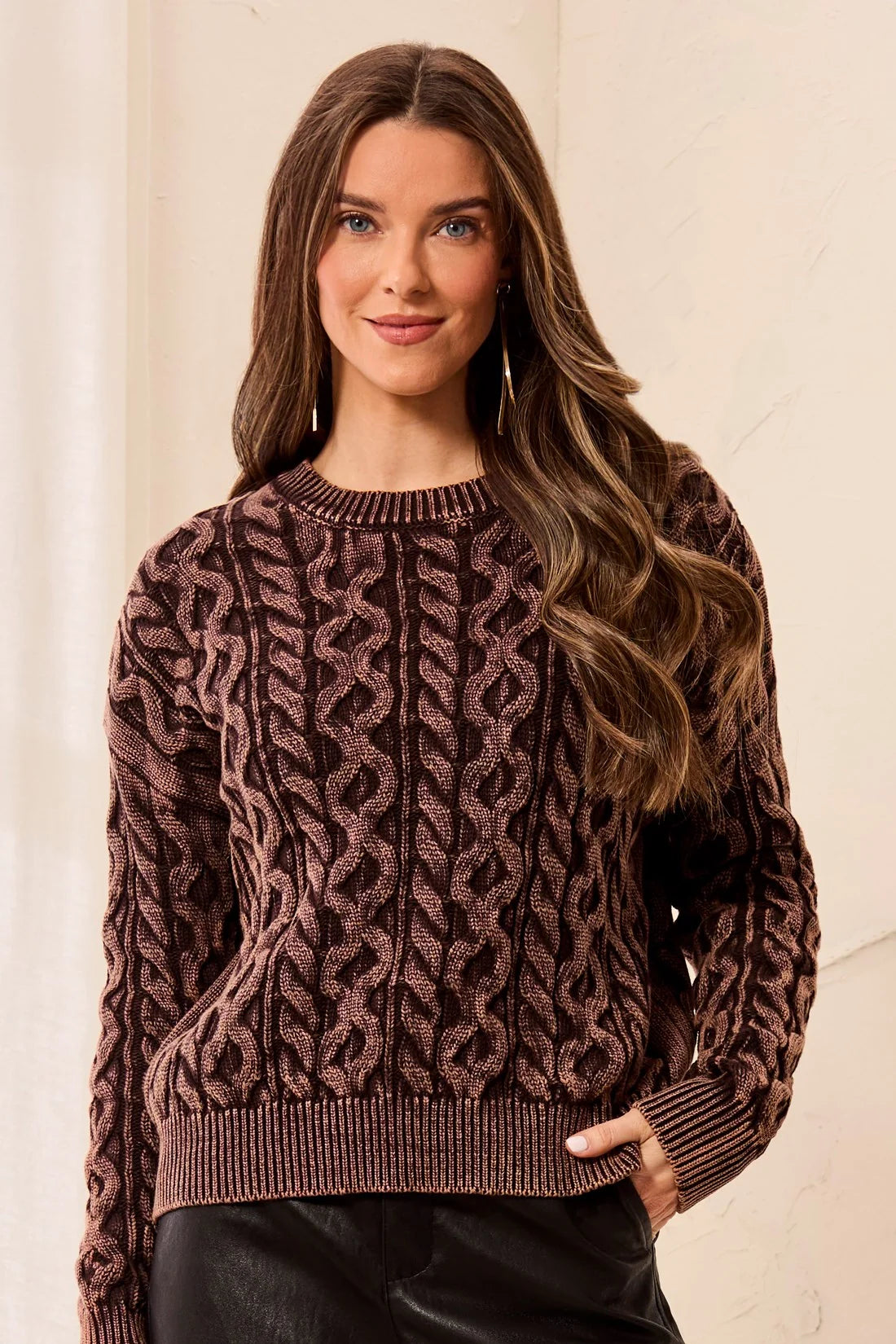 A woman wearing a brown and black cable-knit crewneck sweater, standing against a light beige wall.