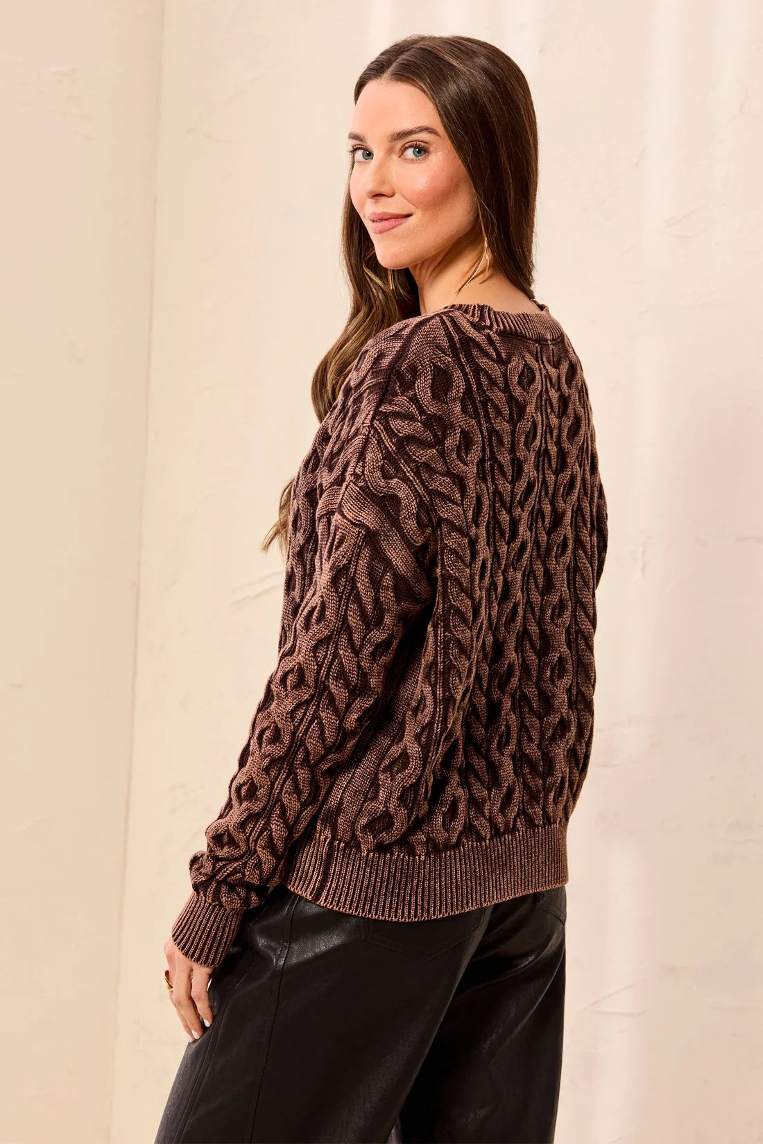 A woman wearing a brown cable-knit sweater with a textured pattern, shown from a side-back angle against a light beige wall.