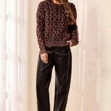 A woman modeling a brown and black patterned knit sweater paired with black pants and black ankle boots, standing in a brightly lit indoor setting with soft curtains.