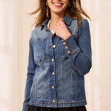 A woman wearing a medium-wash blue denim jacket with button-front closure and chest pockets, paired with black pants, standing against a light cream background.
