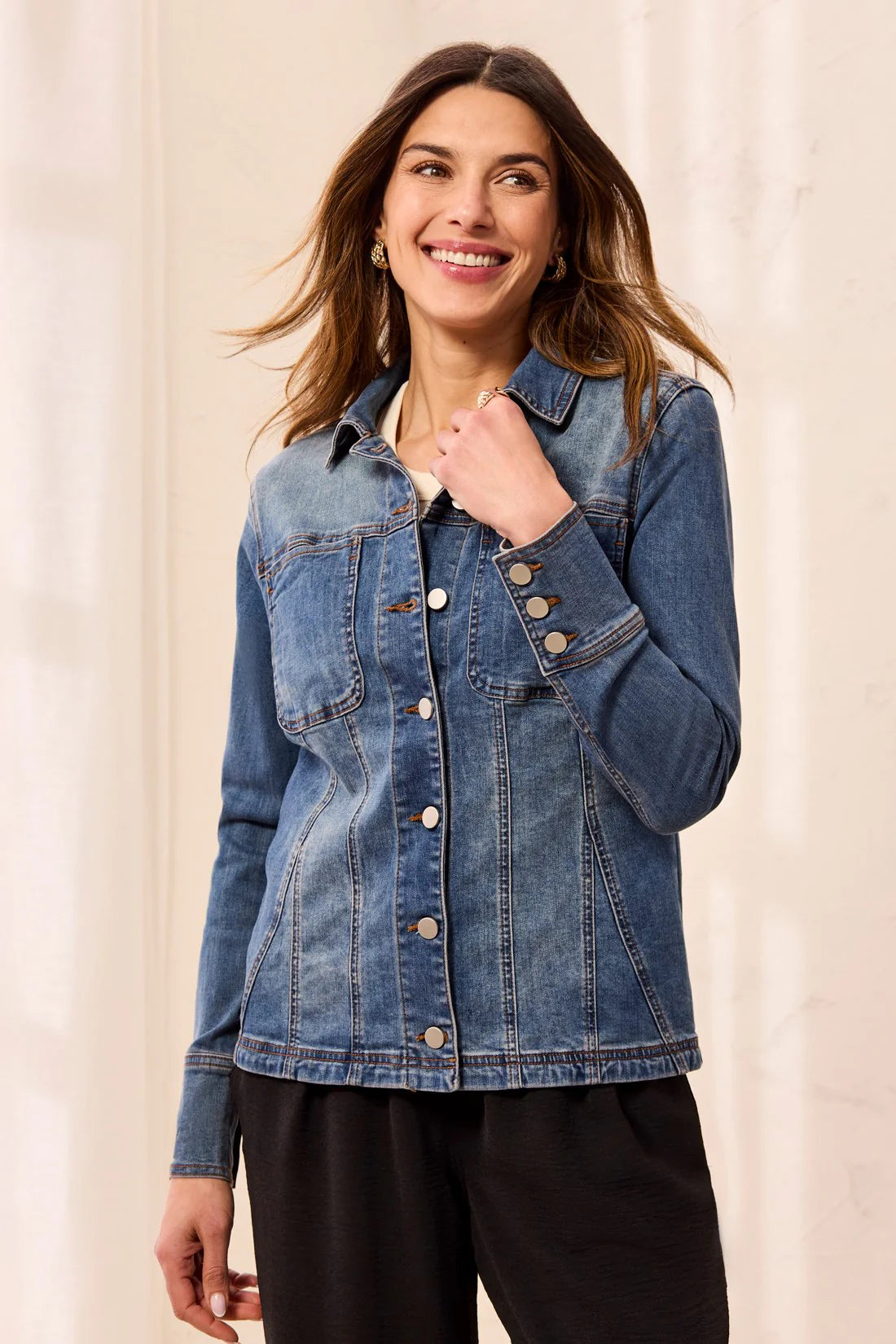 A woman wearing a medium-wash blue denim jacket with button-front closure and chest pockets, paired with black pants, standing against a light cream background.