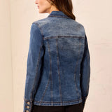 A woman shown from the back wearing a medium wash blue denim jacket with button details on the cuffs, paired with black pants, against a light beige textured wall.