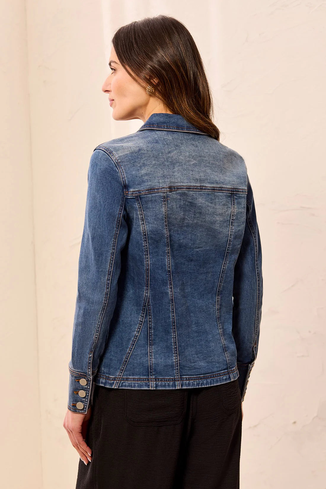 A woman shown from the back wearing a medium wash blue denim jacket with button details on the cuffs, paired with black pants, against a light beige textured wall.