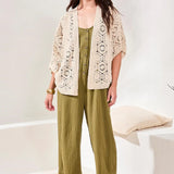 Woman wearing a beige crochet cardigan over a green jumpsuit with wide-leg pants in a minimal indoor setting.