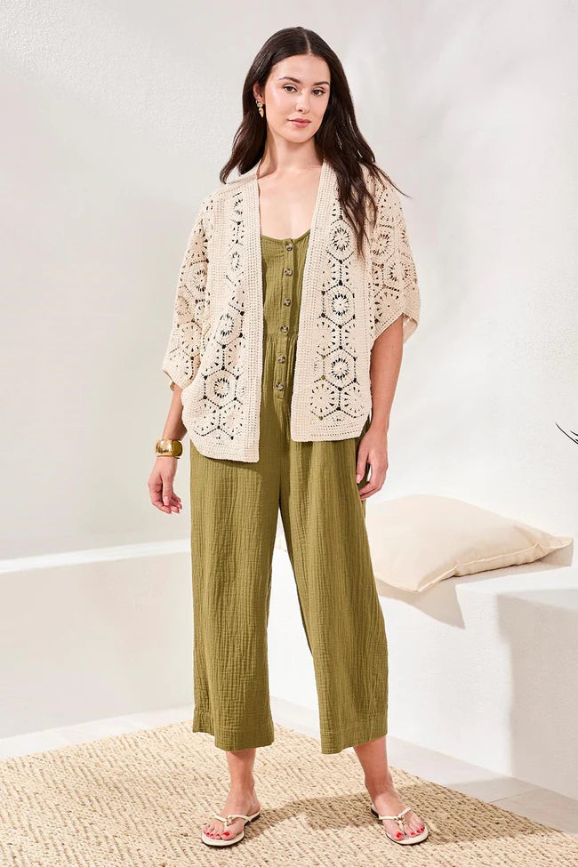 Woman wearing a beige crochet cardigan over a green jumpsuit with wide-leg pants in a minimal indoor setting.