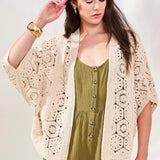 Woman wearing a beige crochet cardigan over a green jumpsuit on a light background