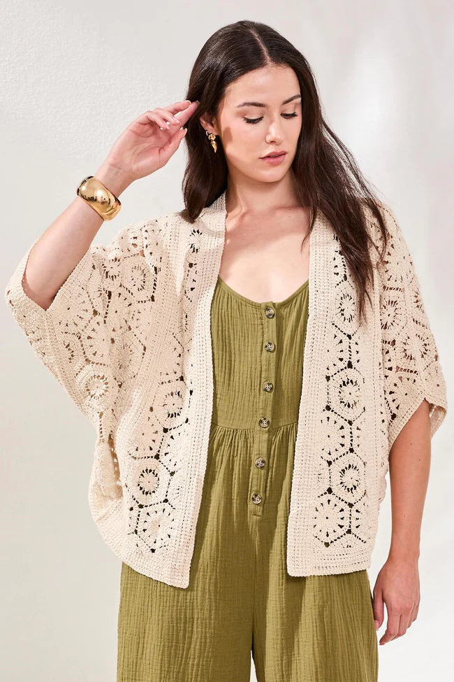 Woman wearing a beige crochet cardigan over a green jumpsuit on a light background