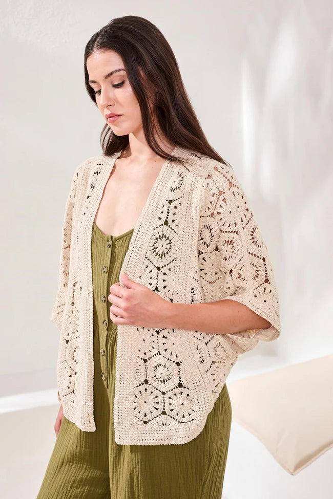 Woman wearing a beige crochet cardigan over a green jumpsuit against a white background