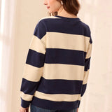 A woman wearing a navy blue and cream striped long-sleeve sweatshirt, shown from the back. She is wearing blue jeans and standing against a soft beige curtain background.