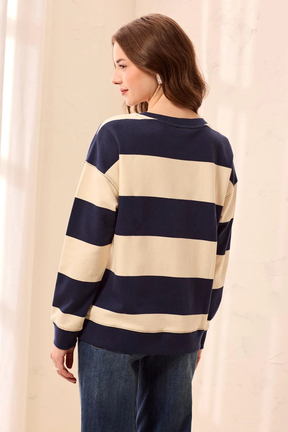 A woman wearing a navy blue and cream striped long-sleeve sweatshirt, shown from the back. She is wearing blue jeans and standing against a soft beige curtain background.