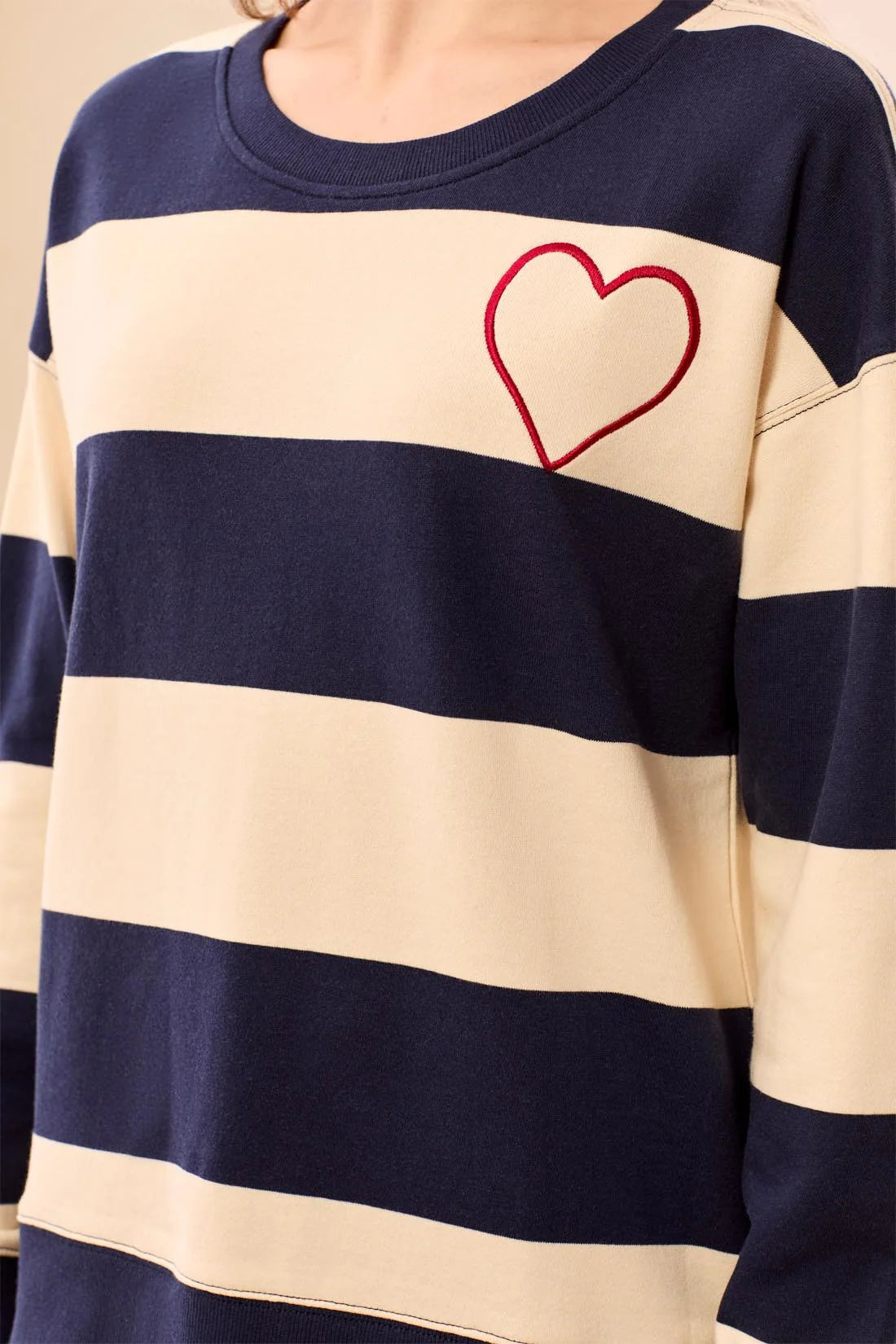 A close-up of a person wearing a long-sleeve crewneck sweatshirt with horizontal navy blue and cream stripes, featuring a red outline heart embroidered on the chest.