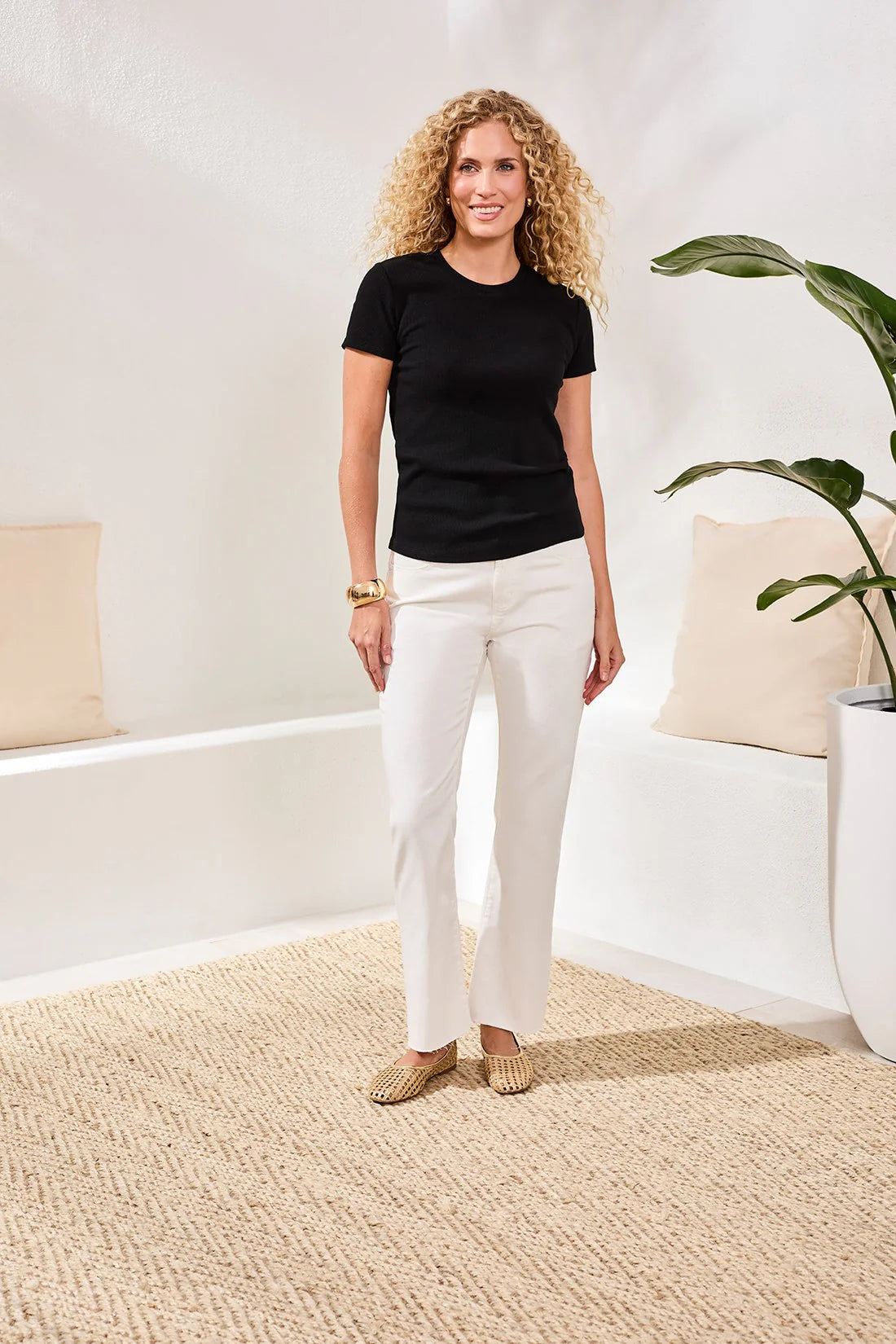 Woman wearing a black t-shirt and white pants standing in a room with a plant and cushions.