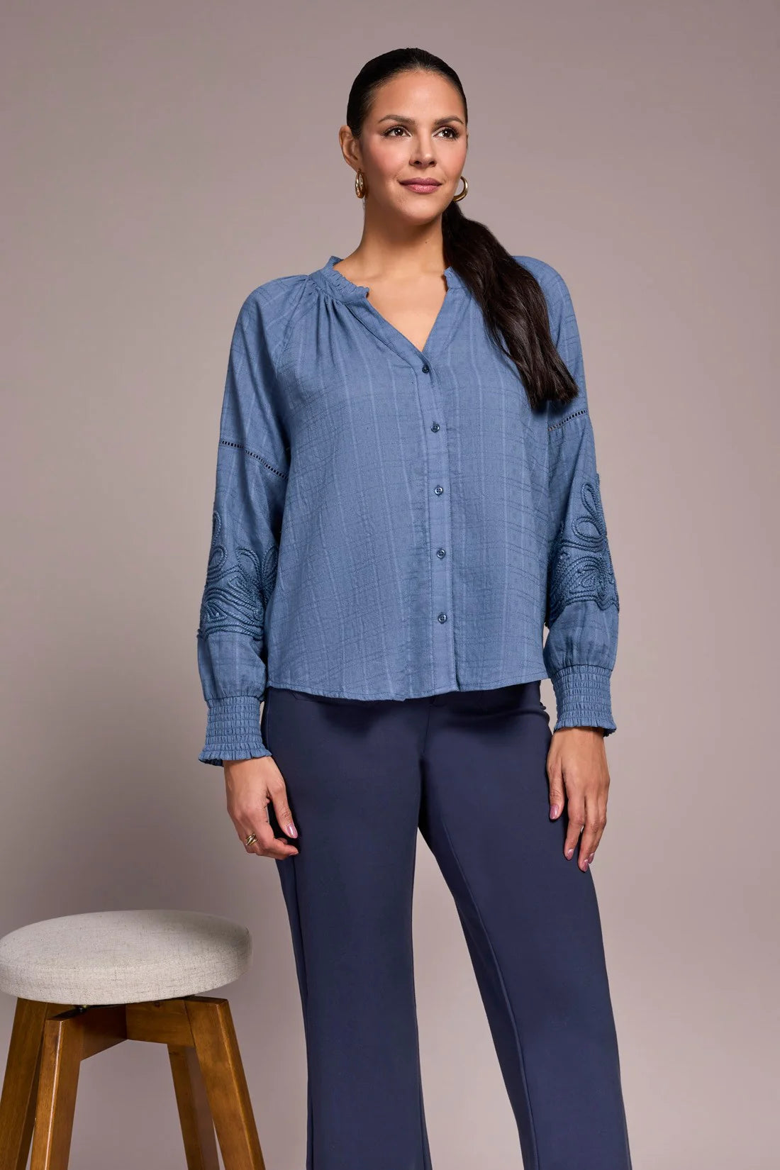 Woman wearing a blue shirt and dark pants standing against a plain background