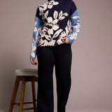 Woman wearing a navy sweater with white floral patterns, standing next to a wooden stool against a plain background.